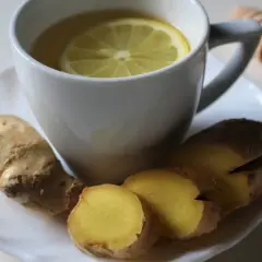 Taza de infusión de jengibre con rodaja de limón y raíz de jengibre fresca. Un palito de miel descansa al lado.