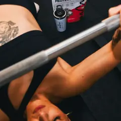 Mujer haciendo press de banca con barra. A su lado, un bote de proteína con leucina, esencial para la recuperación muscular.