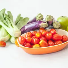 Un surtido vibrante de frutas de temporada: tomates cherry en un cuenco naranja, berenjenas moradas, bok choy verde y dos manzanas verdes brillantes.
