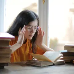 Joven con gafas y libros, con las manos en las sienes, buscando alivio al estrés con magnesio.