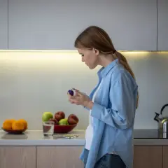 Mujer en cocina revisa frasco, quizás preguntándose inositol que es. Frutas y pastillas en encimera.