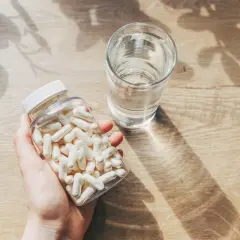 Mano sosteniendo un frasco de cápsulas blancas junto a un vaso de agua. Consulta cuánto zinc tomar al día para tu salud.