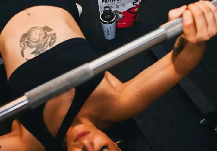 Mujer haciendo press de banca con barra. A su lado, un bote de proteína con leucina, esencial para la recuperación muscular.