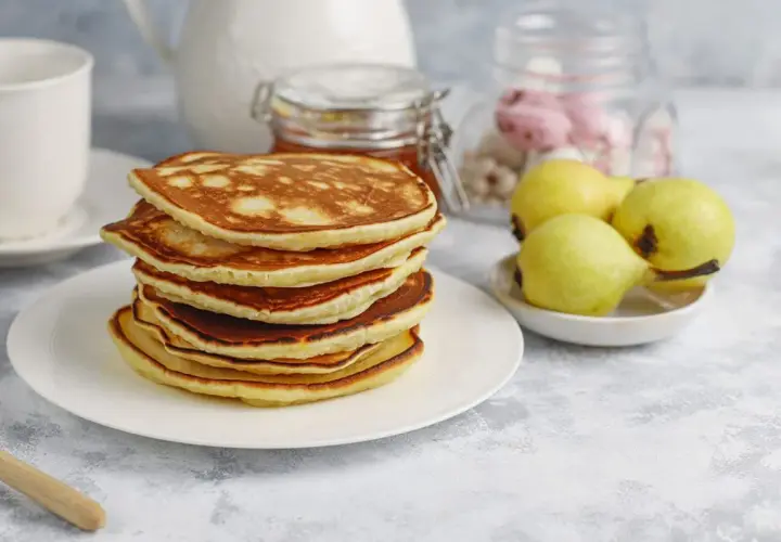 Pila de tortitas fit doradas y esponjosas, acompañadas de peras y un tarro de miel. Un desayuno saludable y delicioso.