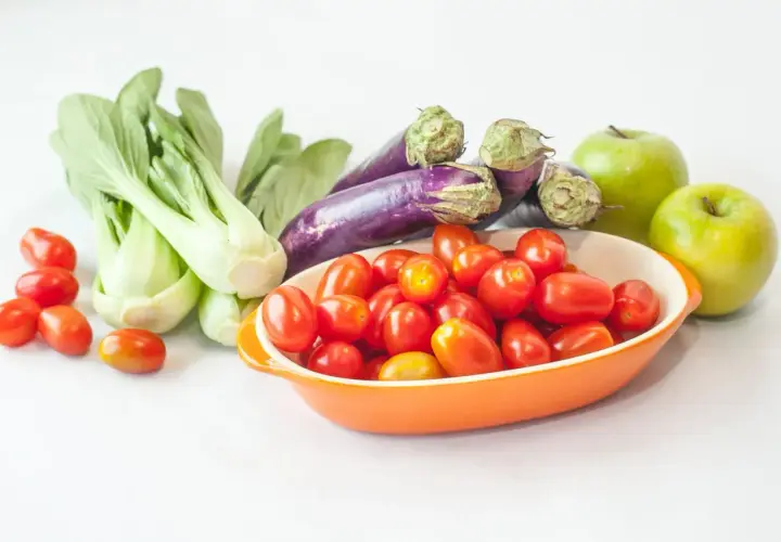 Un surtido vibrante de frutas de temporada: tomates cherry en un cuenco naranja, berenjenas moradas, bok choy verde y dos manzanas verdes brillantes.