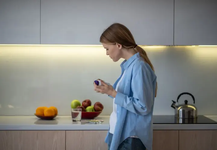 Mujer en cocina revisa frasco, quizás preguntándose inositol que es. Frutas y pastillas en encimera.