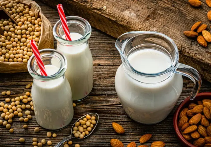 Botellas y jarra con bebida vegetal, junto a un saco de soja y almendras.