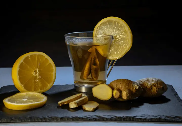 Un vaso de té con limón y jengibre, acompañado de rodajas de limón y trozos de jengibre fresco. Un shot de jengibre para revitalizar.