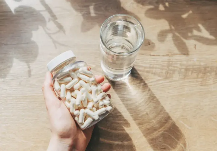 Mano sosteniendo un frasco de cápsulas blancas junto a un vaso de agua. Consulta cuánto zinc tomar al día para tu salud.