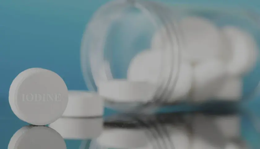 Pastillas de yodo blancas, una en primer plano con la palabra "IODINE" grabada, y un frasco volcado con más pastillas.