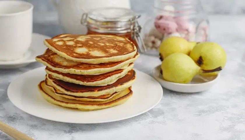 Pila de tortitas fit doradas y esponjosas, acompañadas de peras y un tarro de miel. Un desayuno saludable y delicioso.