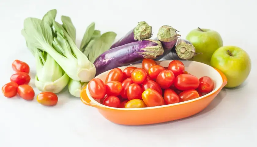 Un surtido vibrante de frutas de temporada: tomates cherry en un cuenco naranja, berenjenas moradas, bok choy verde y dos manzanas verdes brillantes.
