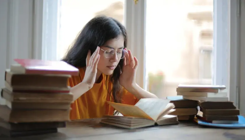 Joven con gafas y libros, con las manos en las sienes, buscando alivio al estrés con magnesio.