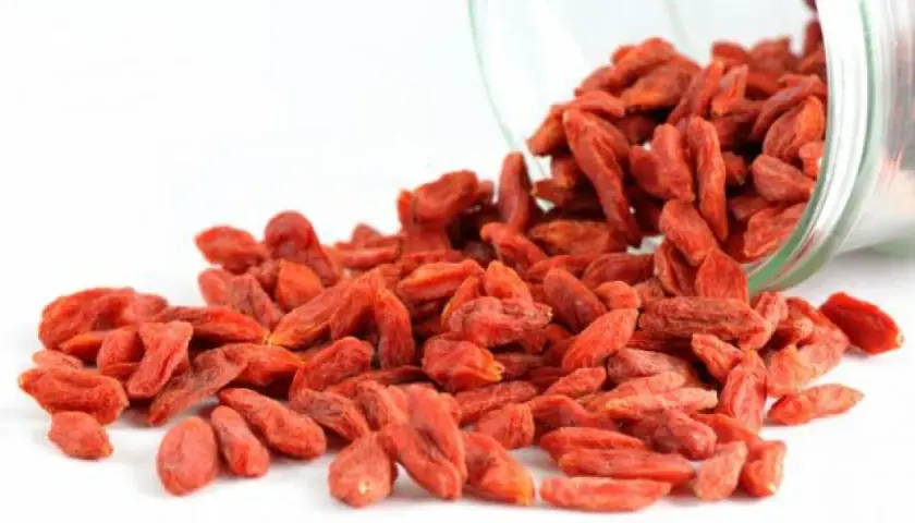 Un frasco de vidrio derrama bayas de goji, destacando sus vibrantes bayas rojas y sus innumerables propiedades beneficiosas.