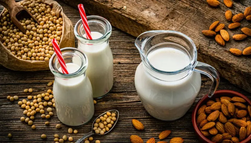 Botellas y jarra con bebida vegetal, junto a un saco de soja y almendras.