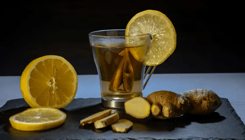 Un vaso de té con limón y jengibre, acompañado de rodajas de limón y trozos de jengibre fresco. Un shot de jengibre para revitalizar.