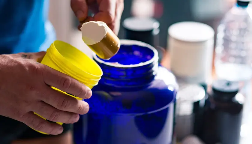 Man prepara su batido de creatina, iniciando la fase de carga con polvo blanco en un gran recipiente azul.