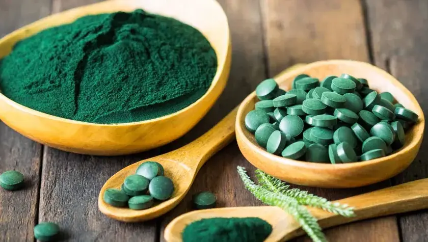 Polvo y pastillas verdes vibrantes, que muestran qué es la chlorella, en cuencos de madera y cucharas sobre una superficie rústica.