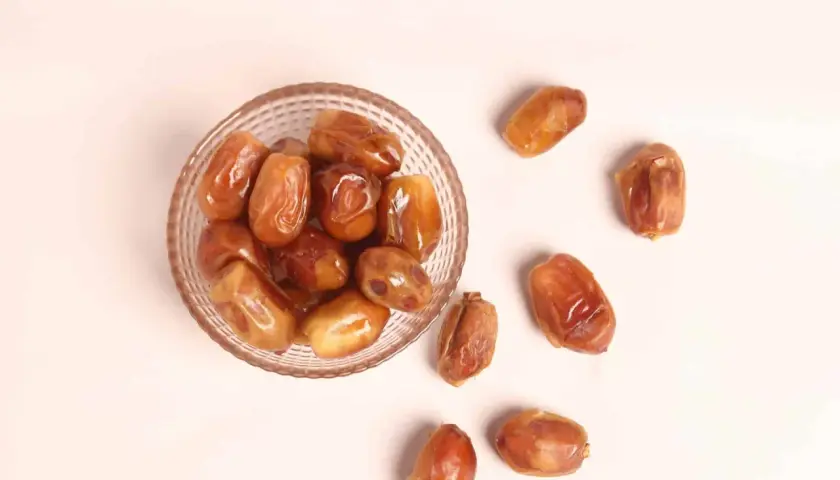 Un cuenco de cristal lleno de dátiles jugosos y otros esparcidos alrededor, destacando sus nutritivas propiedades.