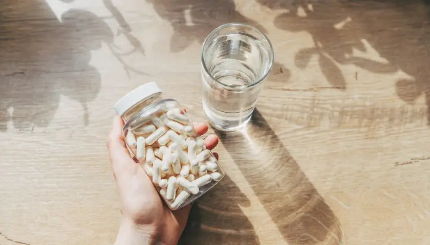 Mano sosteniendo un frasco de cápsulas blancas junto a un vaso de agua. Consulta cuánto zinc tomar al día para tu salud.
