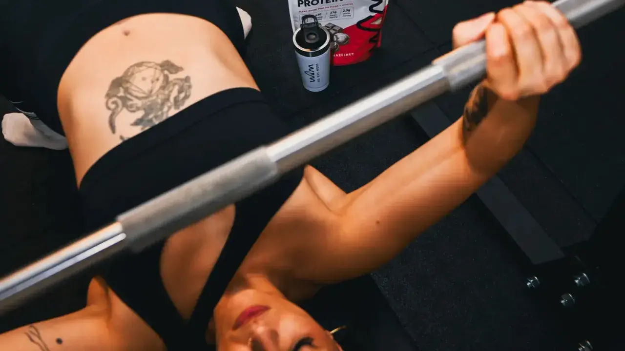 Mujer haciendo press de banca con barra. A su lado, un bote de proteína con leucina, esencial para la recuperación muscular.
