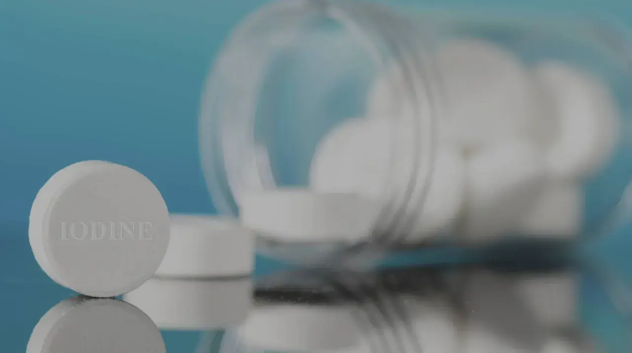 Pastillas de yodo blancas, una en primer plano con la palabra "IODINE" grabada, y un frasco volcado con más pastillas.