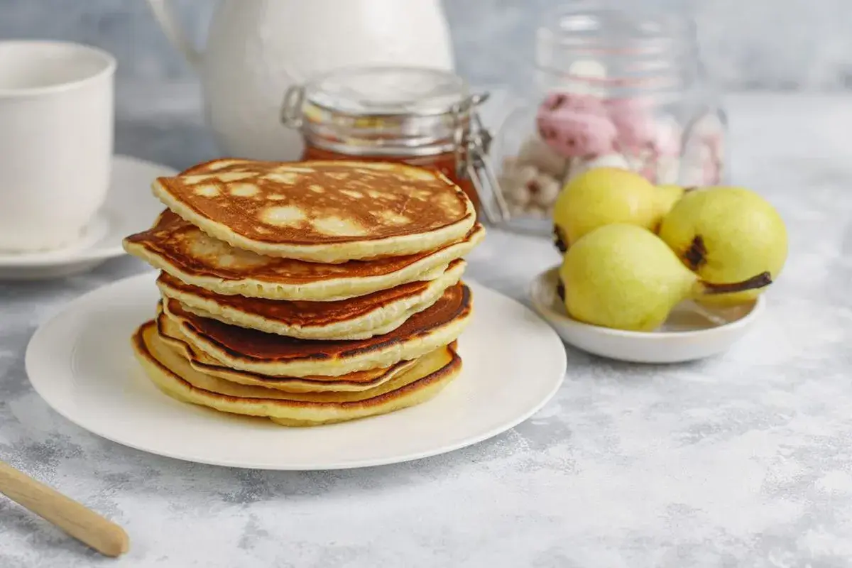 Pila de tortitas fit doradas y esponjosas, acompañadas de peras y un tarro de miel. Un desayuno saludable y delicioso.
