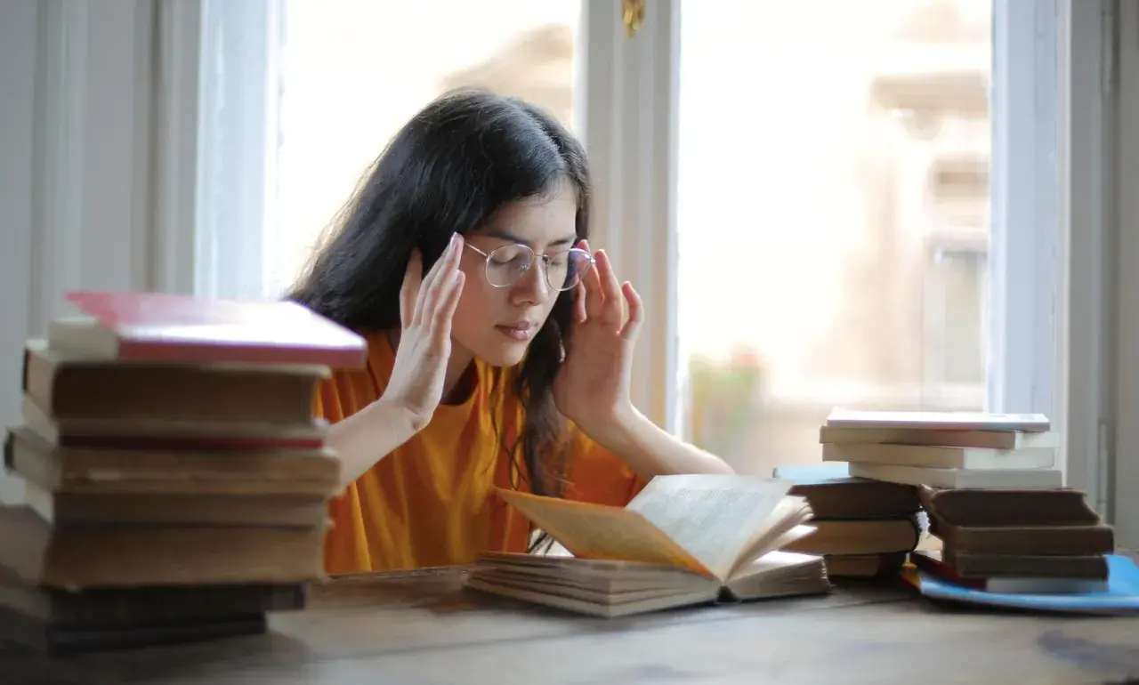 Joven con gafas y libros, con las manos en las sienes, buscando alivio al estrés con magnesio.
