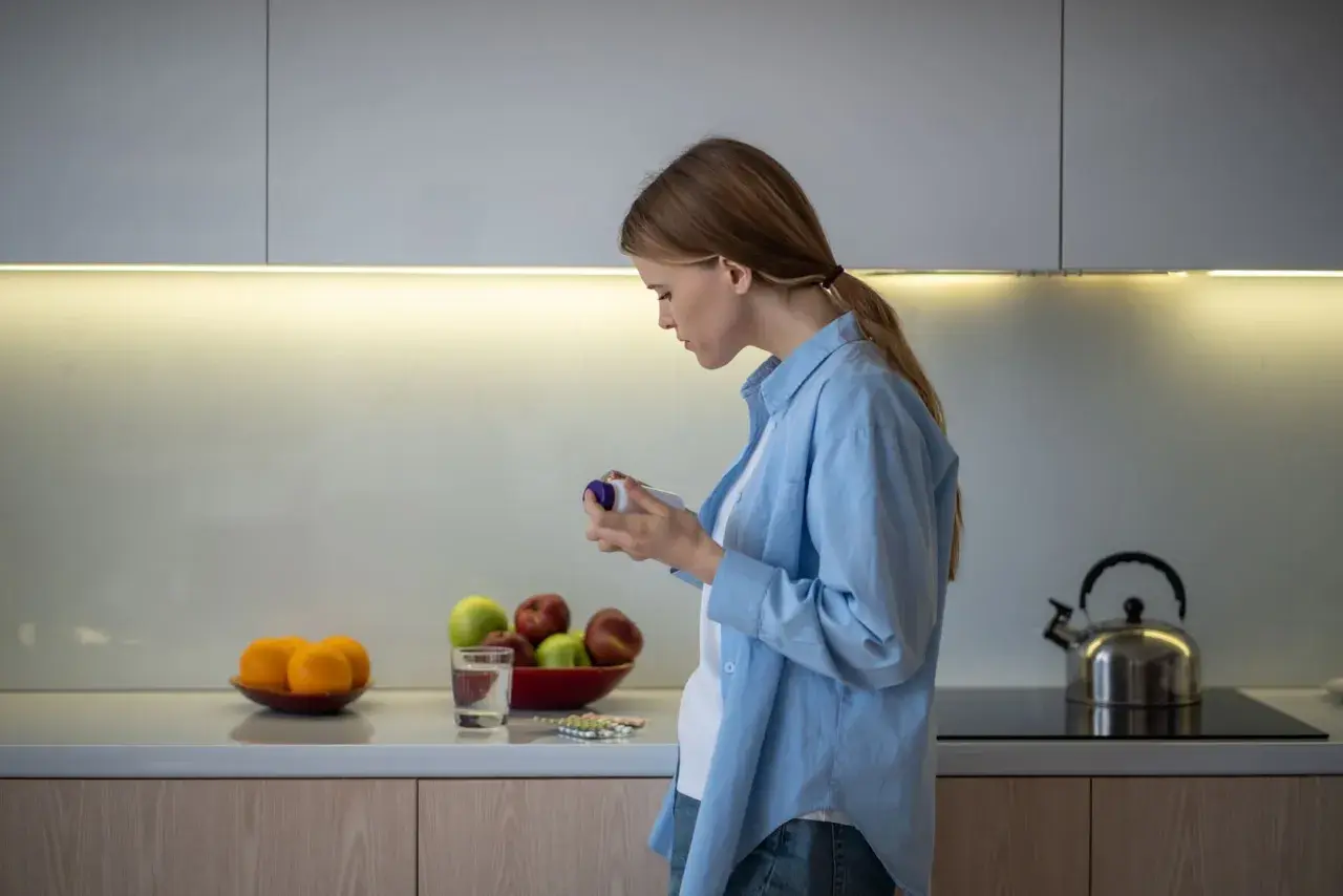 Mujer en cocina revisa frasco, quizás preguntándose inositol que es. Frutas y pastillas en encimera.