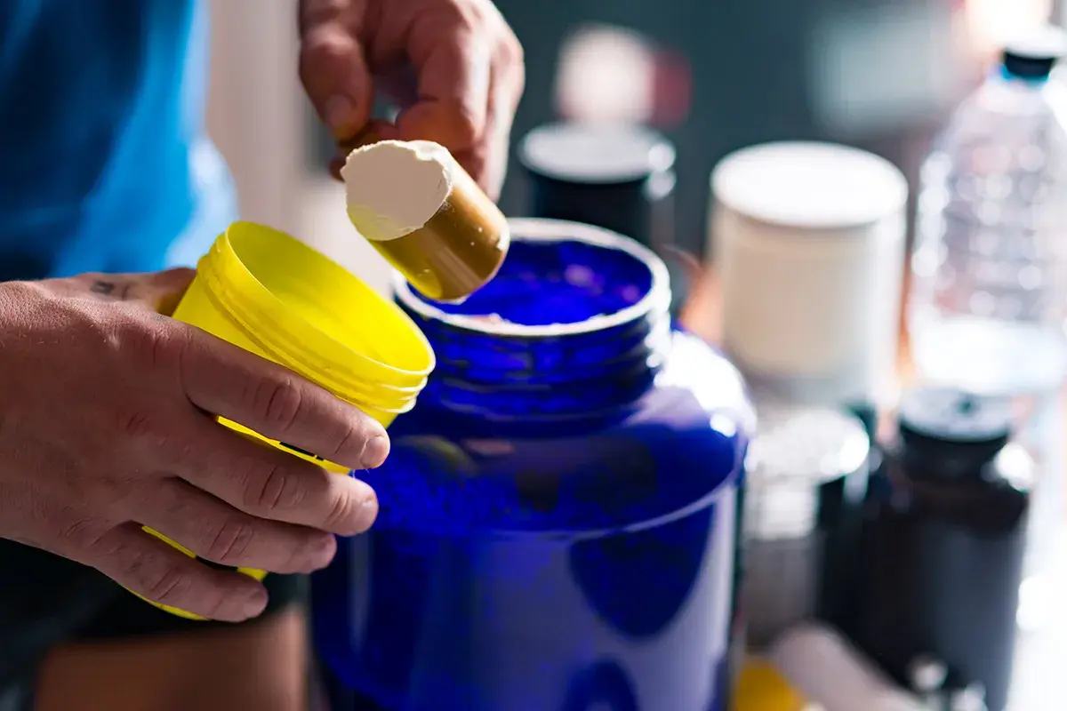 Man prepara su batido de creatina, iniciando la fase de carga con polvo blanco en un gran recipiente azul.