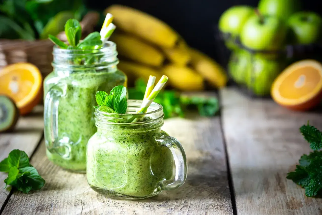 Batidos verdes refrescantes con espirulina, que es un superalimento, adornados con menta. Plátanos, naranjas y manzanas verdes en el fondo.