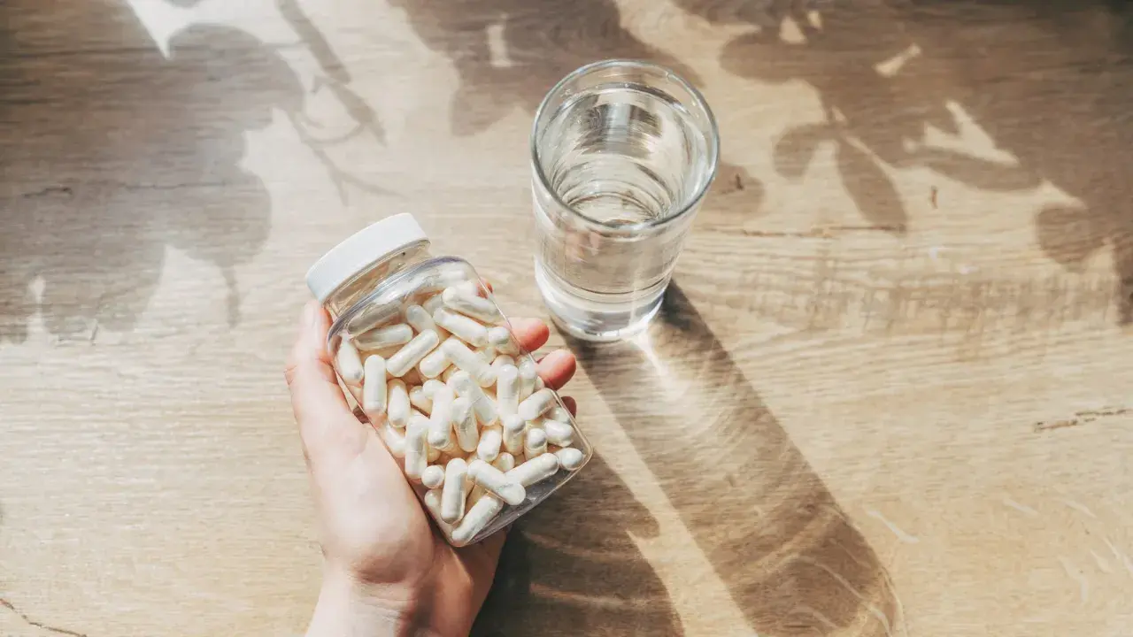 Mano sosteniendo un frasco de cápsulas blancas junto a un vaso de agua. Consulta cuánto zinc tomar al día para tu salud.