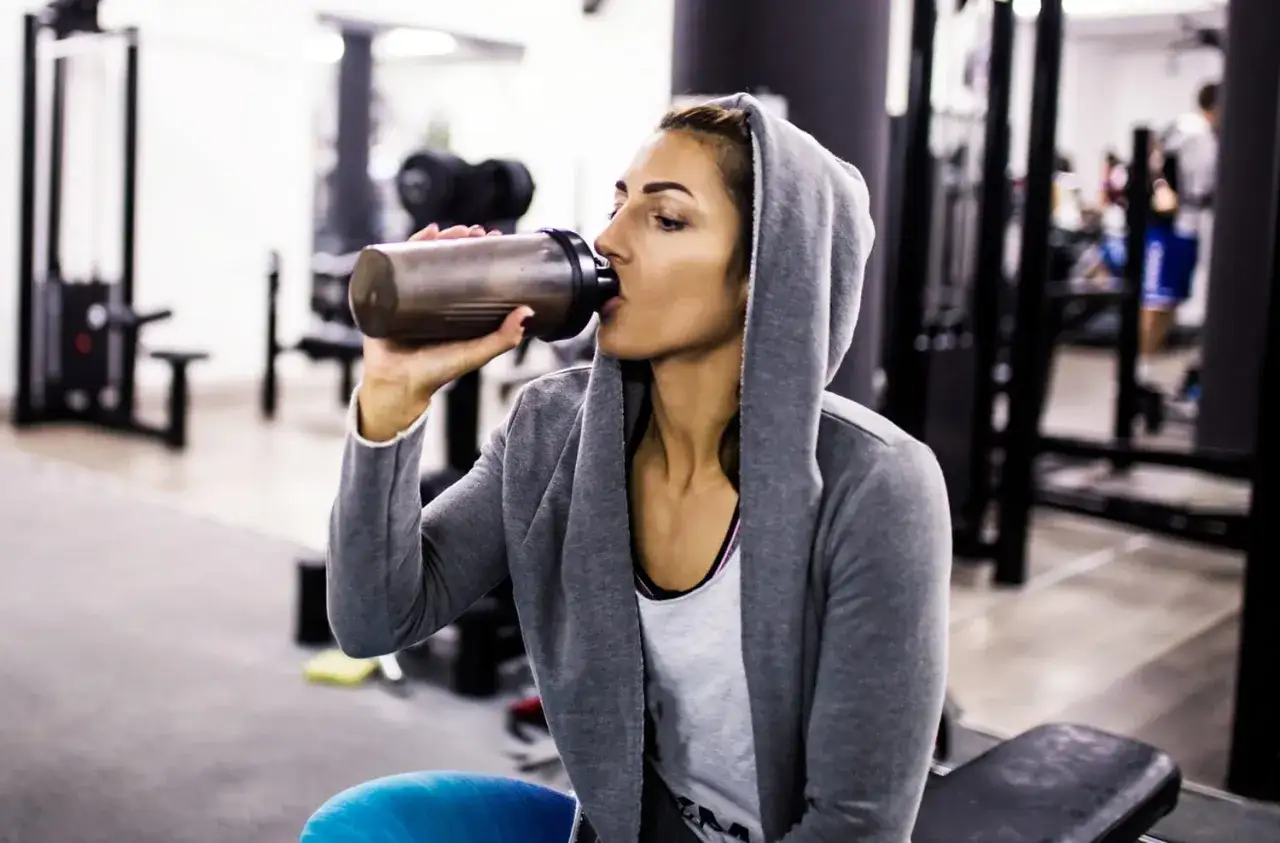 Mujer en el gimnasio bebiendo su batido de proteína, esencial para la recuperación muscular y el bienestar femenino.