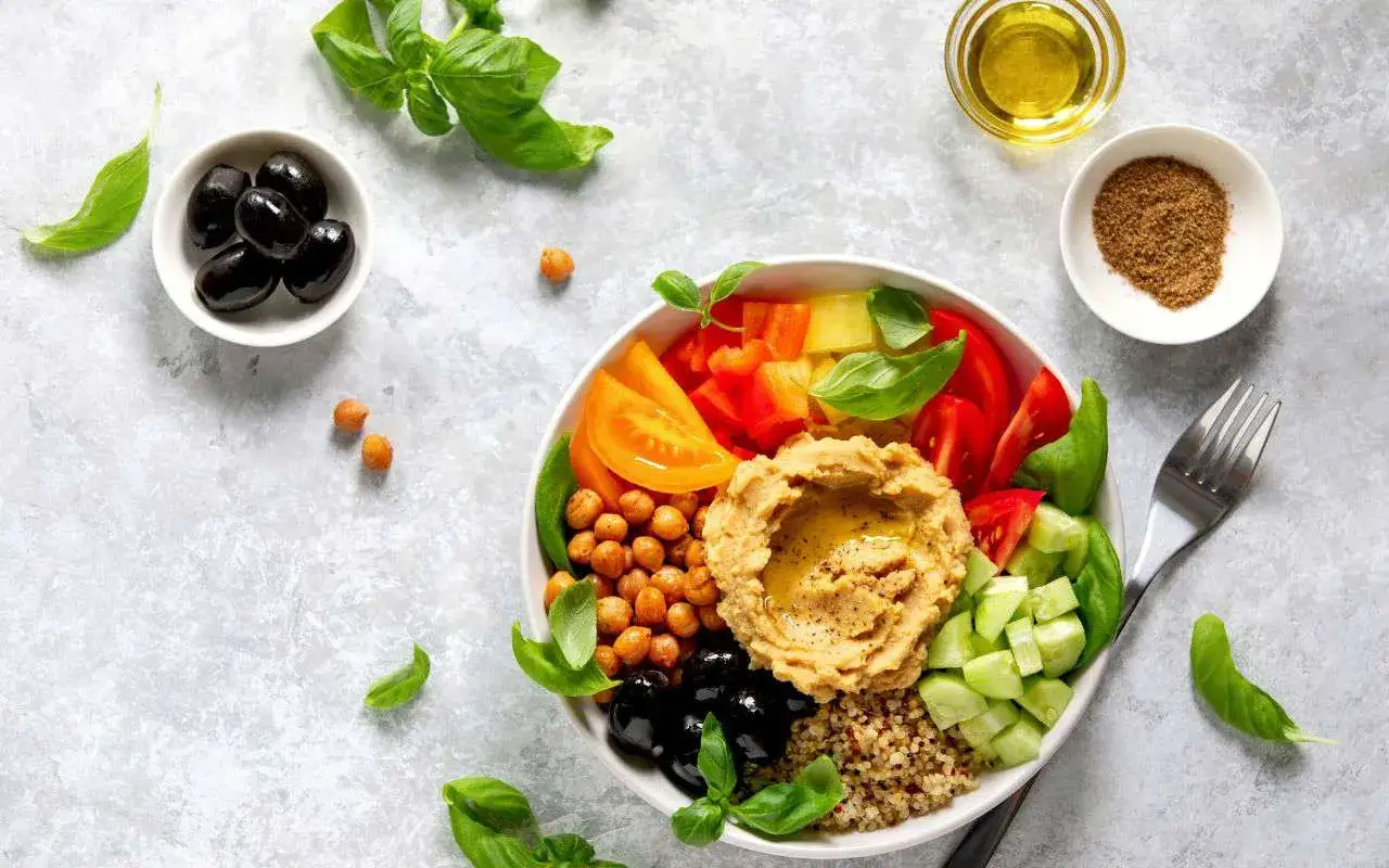 Un bol con garbanzos, aceitunas negras, pepino, tomate, pimientos y hummus. Podría ser un ejemplo de alimentos prohibidos para la colitis ulcerosa.