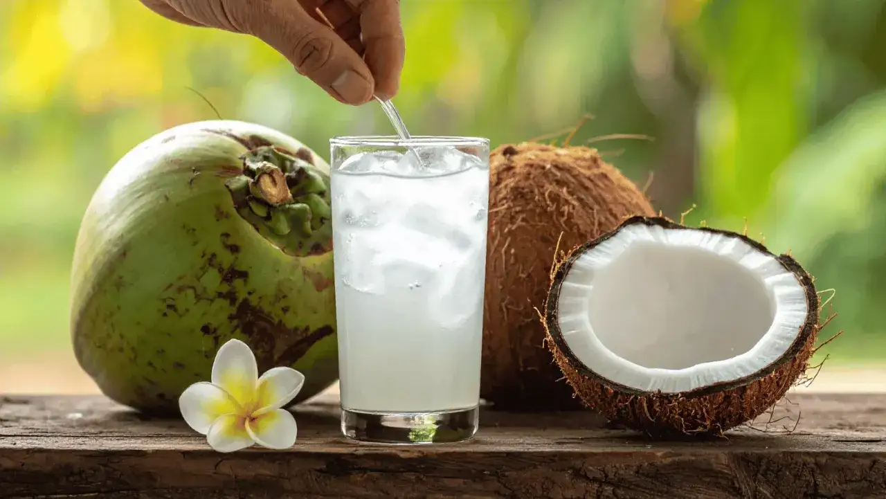 Un vaso de agua de coco refrescante con hielo, junto a cocos enteros y partidos, y una flor tropical. Descubre los agua de coco beneficios.
