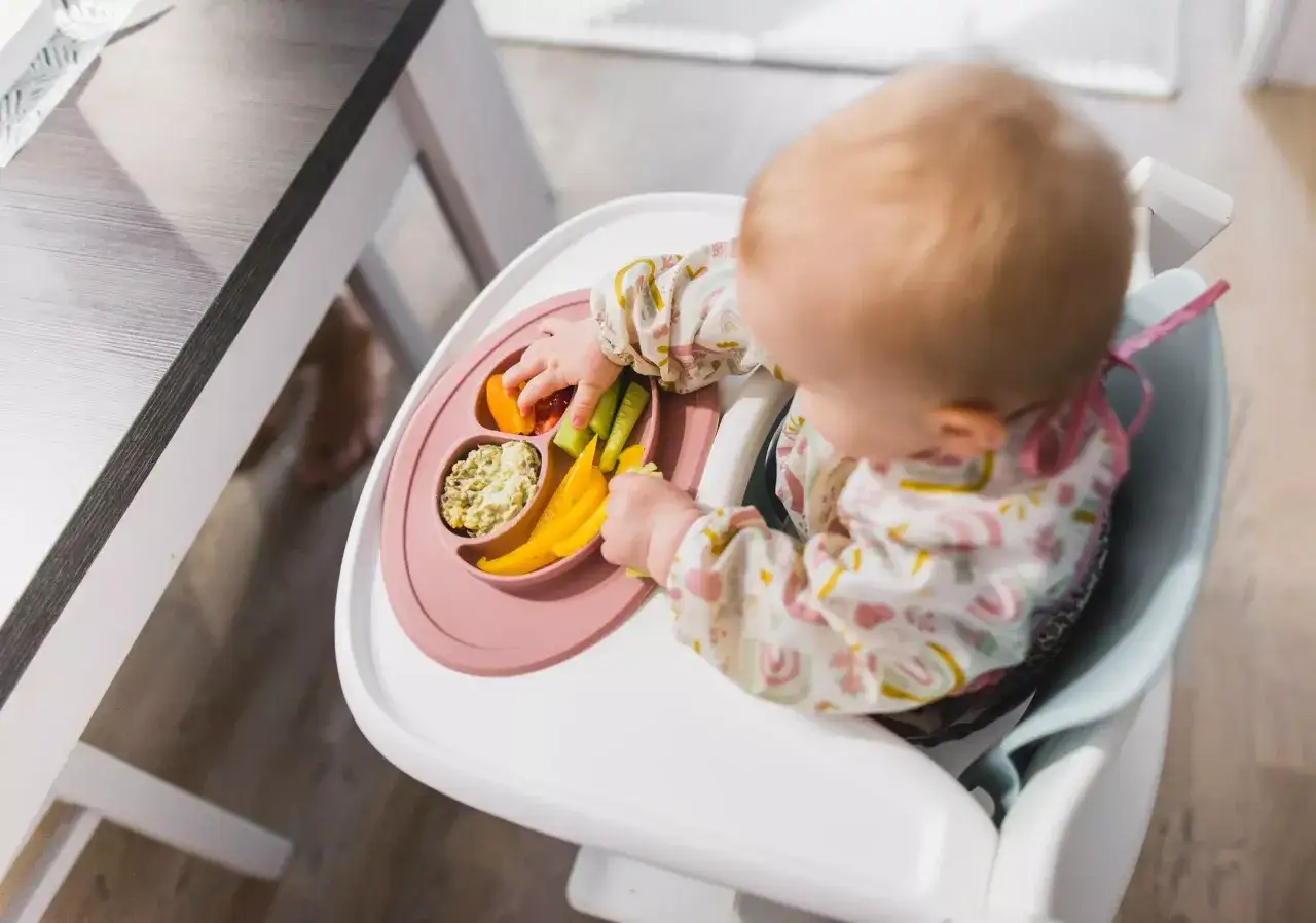 Un bebé explora su comida con el método blw. En su trona, tiene un plato con trozos de pimiento, pepino y mango.