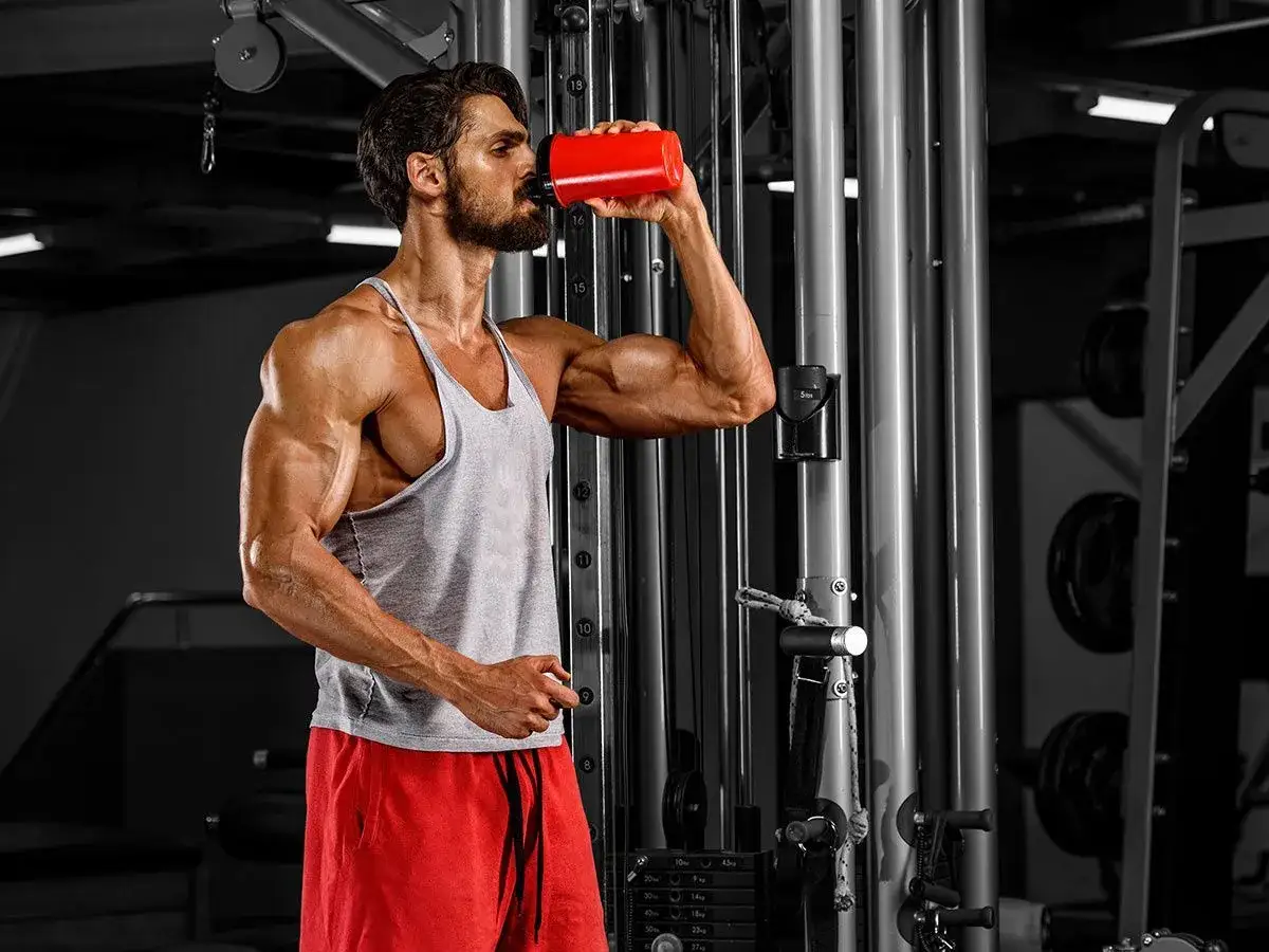 Hombre musculoso bebe de un bid&oacute;n rojo en el gimnasio, un ejemplo de como tomar prote&iacute;na para recuperarse.