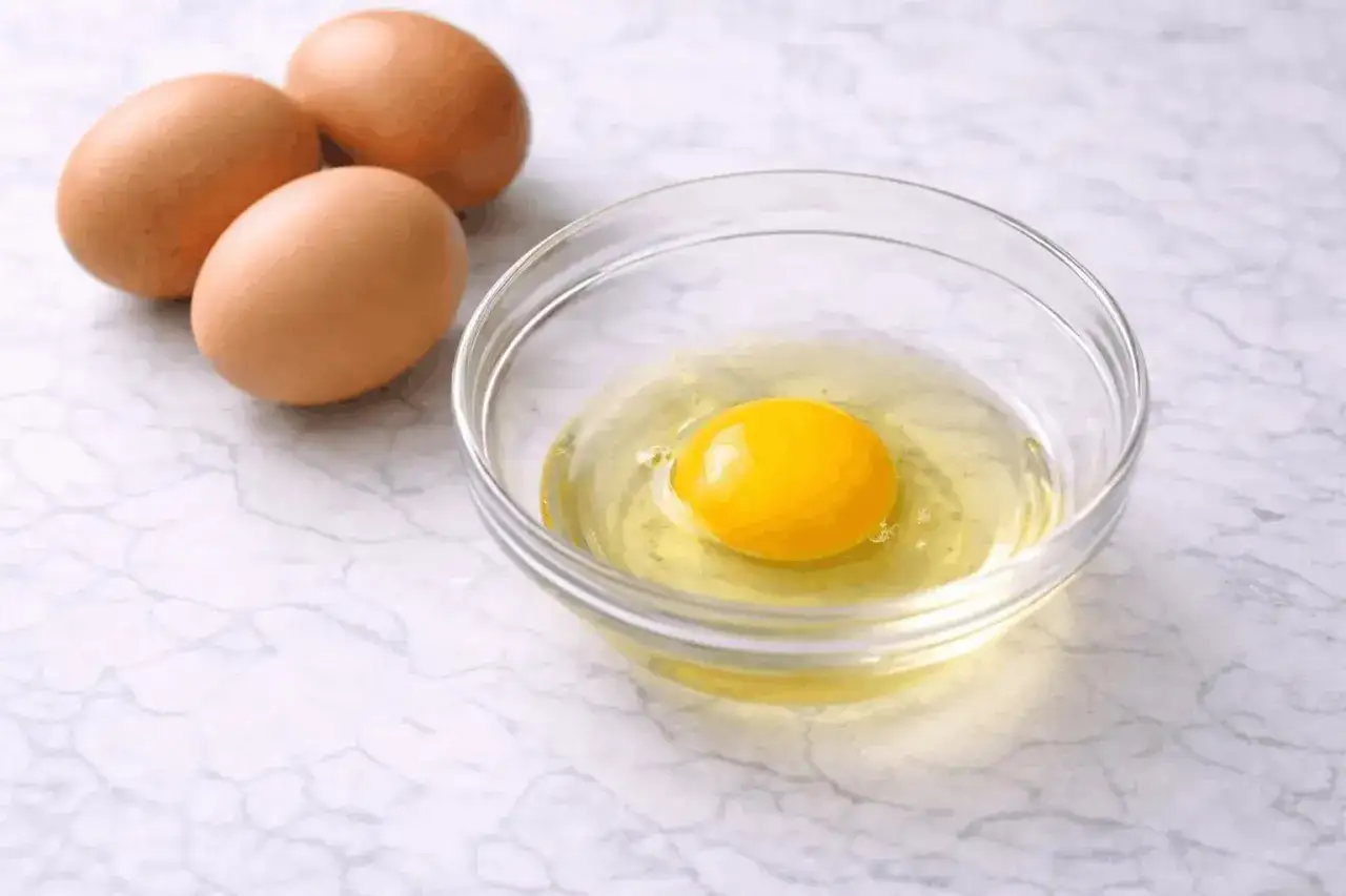 Un huevo crudo con su yema brillante en un bol de cristal, junto a otros dos huevos enteros.