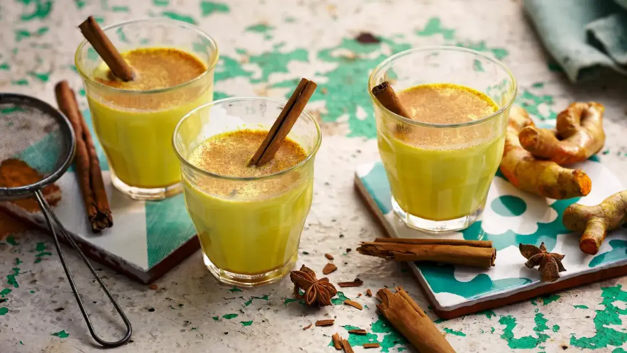 Tres vasos de leche dorada con canela y an&iacute;s estrellado, junto a ra&iacute;ces de c&uacute;rcuma fresca.