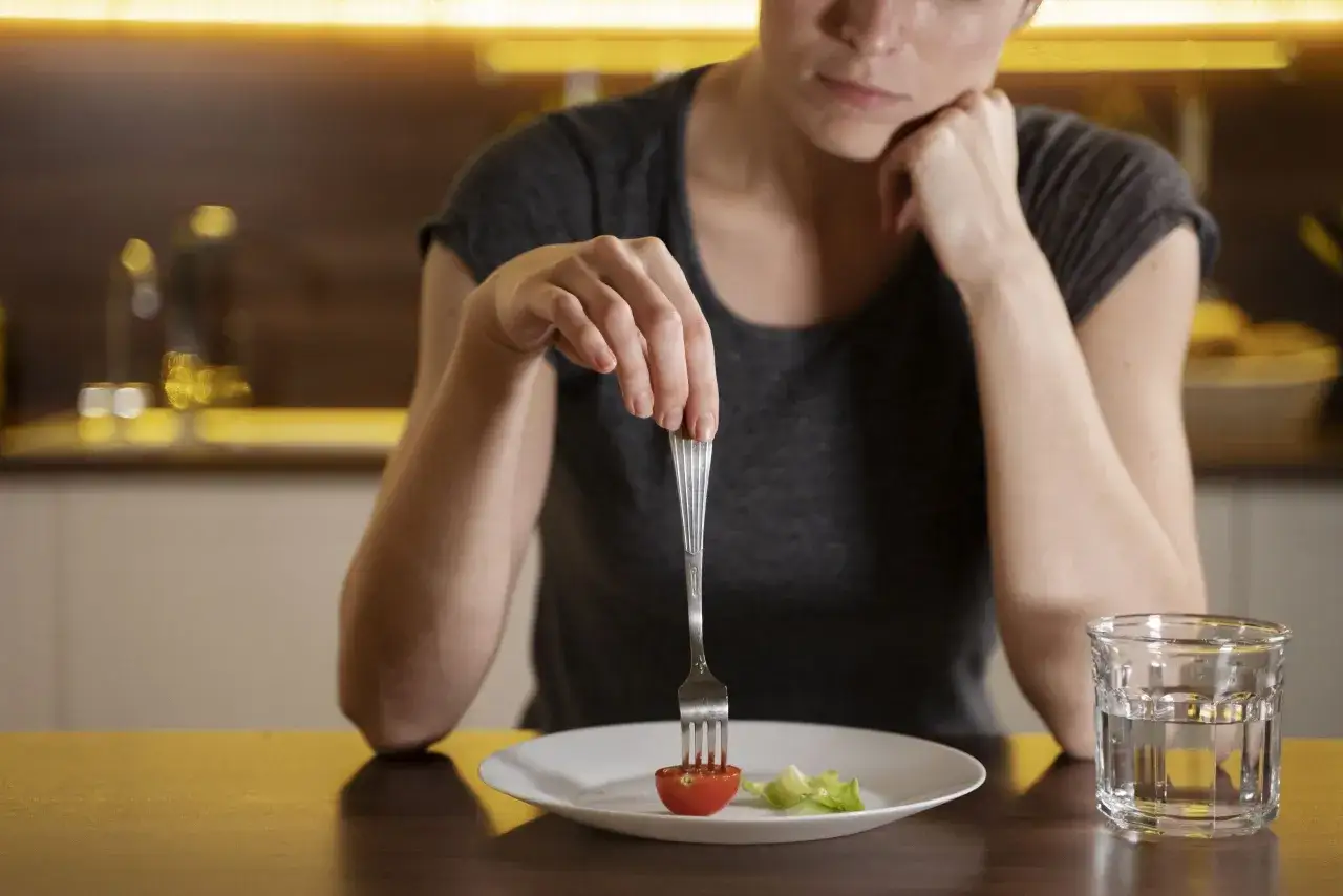 Mujer con expresión de cansancio, con un tenedor clavado en un tomate y una hoja de lechuga en un plato. Un vaso de agua al lado. Parece que está probando dietas milagro.