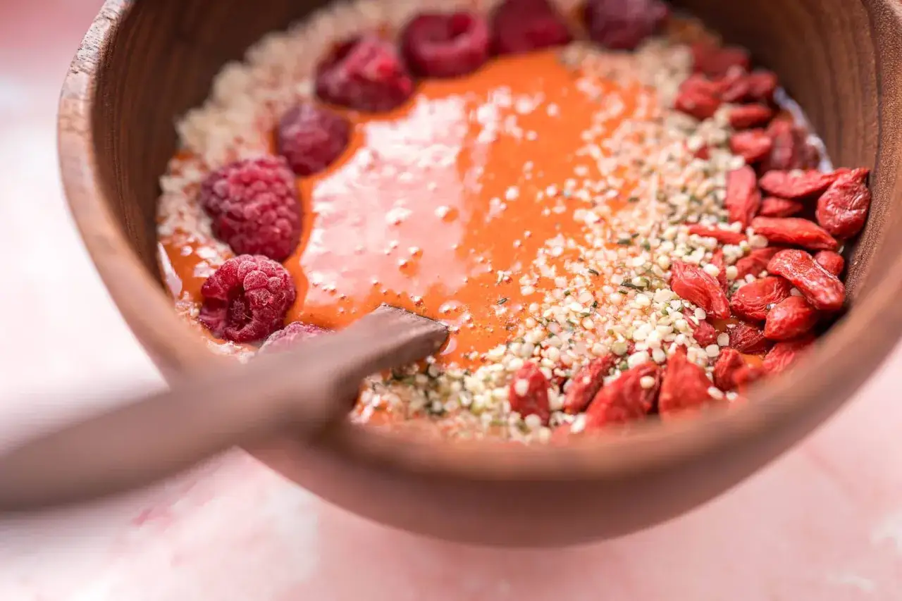 Un tazón de madera con un batido naranja, frambuesas, semillas de cáñamo y bayas de goji, destacando sus propiedades nutritivas.