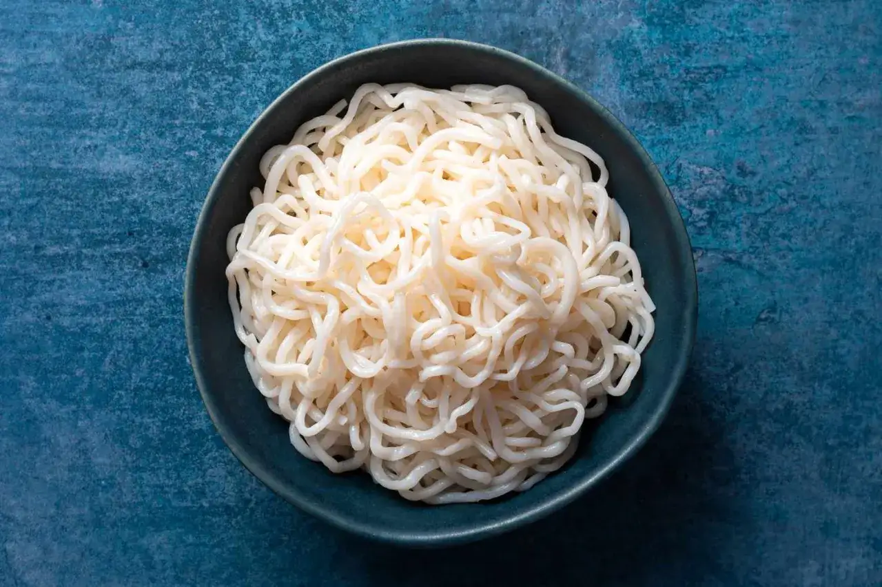 Un tazón de fideos blancos y translúcidos, que es konjac, sobre un fondo azul texturizado.